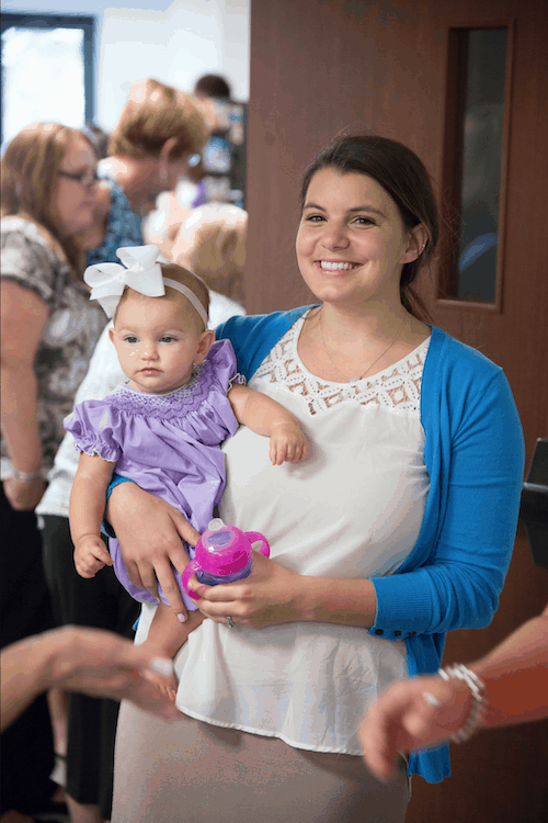 Woman in blue cardigan, smiling and holding her baby girl who is wearing a dress and hairbow.
