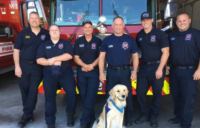 Triton the comfortdog sitting with Fire Department crew in front of their fire truck parked in garage