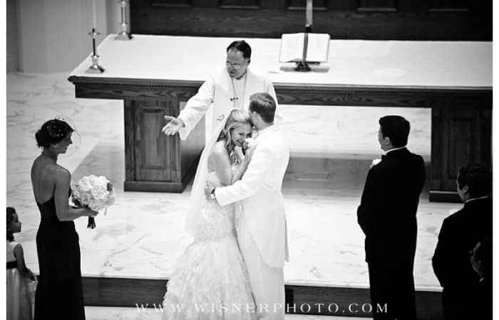Image of bride and groom embracing at the altar with pastor smiling. Watermark of www.wisnerphoto.com